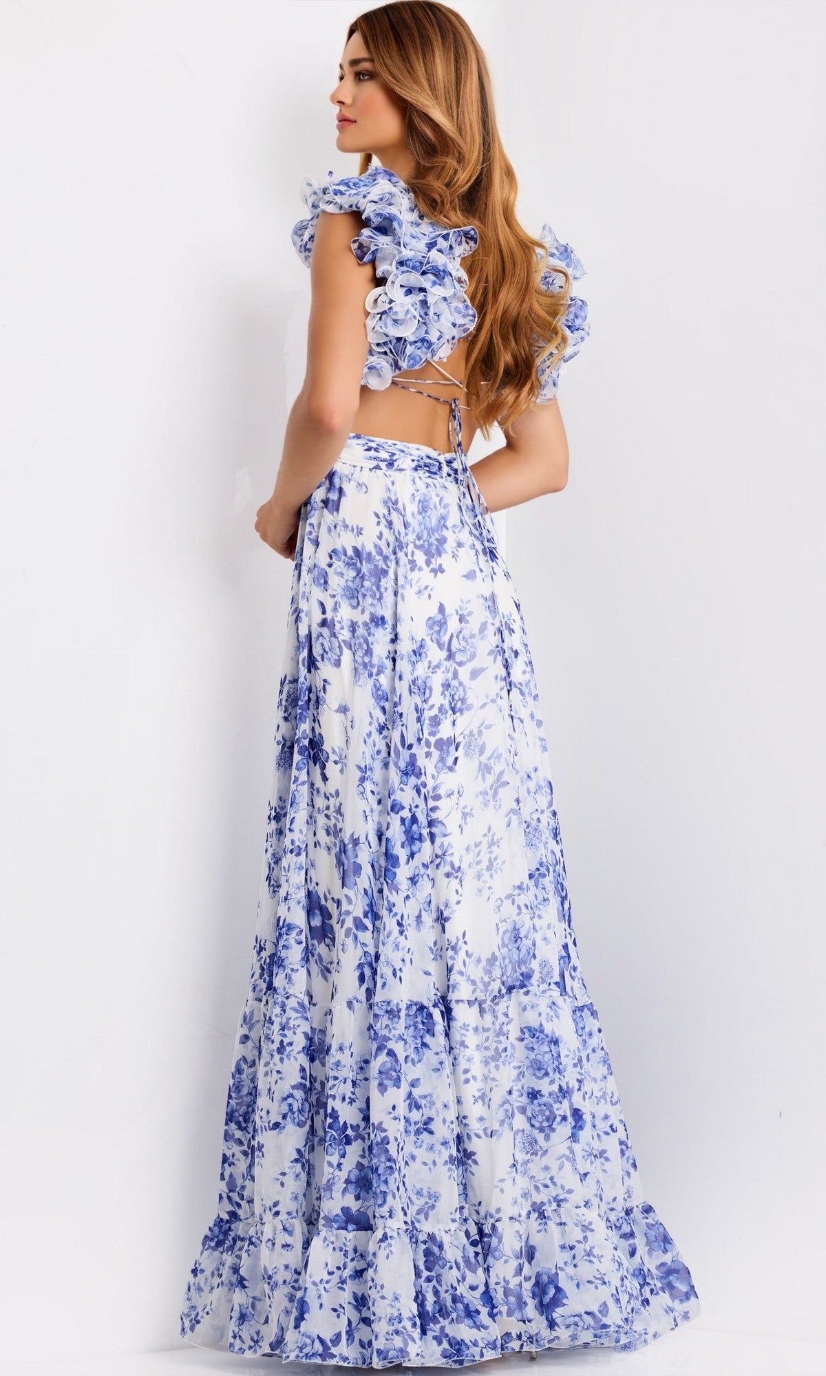 A woman stands sideways in the Jovani 49143 Ruffled Long Print Prom Dress, featuring white and blue florals, ruffled sleeves, and an open back, against a plain white background.